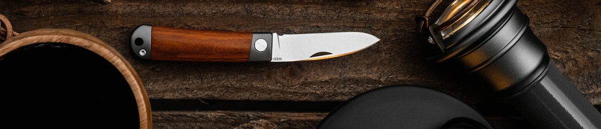 A WESN Henry with cherrywood handle on a wooden table next to a coffee mug, a lantern and a flashlight