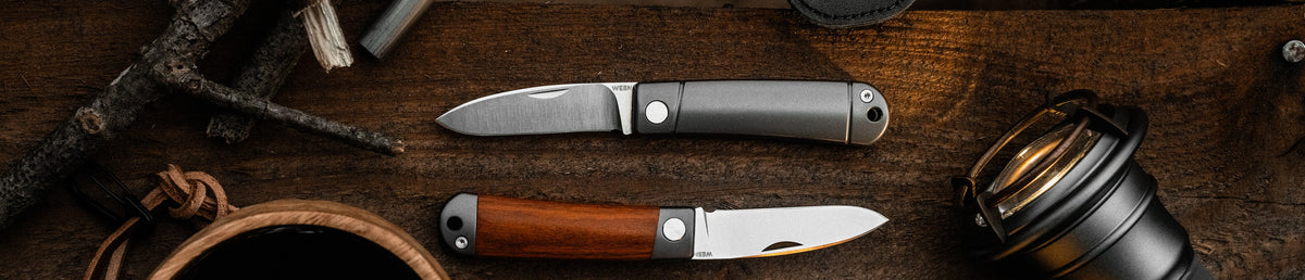Two WESN Henry knives, titanium and cherrywood, on a wooden table beside a coffee mug and a lantern